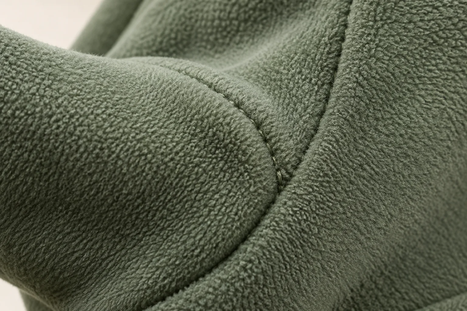 Sage-green fleece glove: macro of smooth soft short pile at palm-thumb curved seam, dense non-knitted fibers and small tonal stitching; no full thumb.