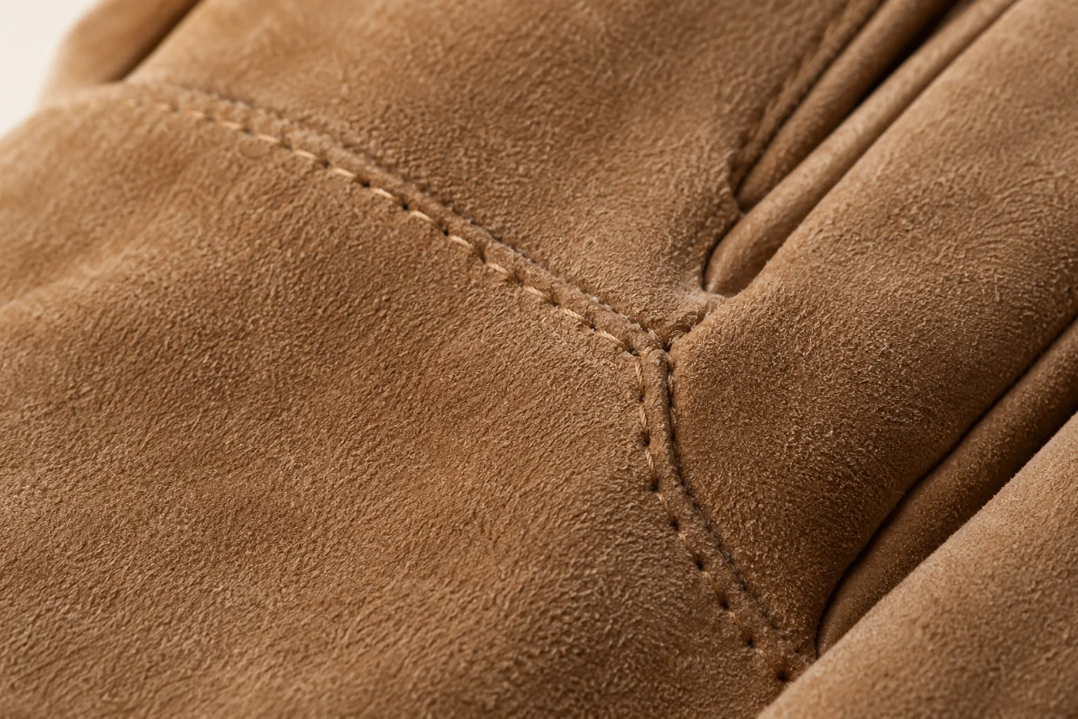 Tan suede winter glove: macro fine matte suede nap at endpoint of a small tonal hand-back stitch line.