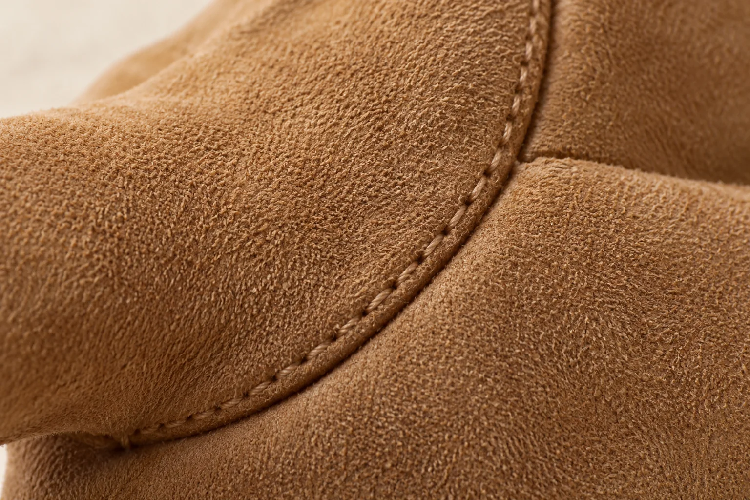 Tan suede winter glove: macro curved thumb-side seam with tan tonal thread joining two suede panels.