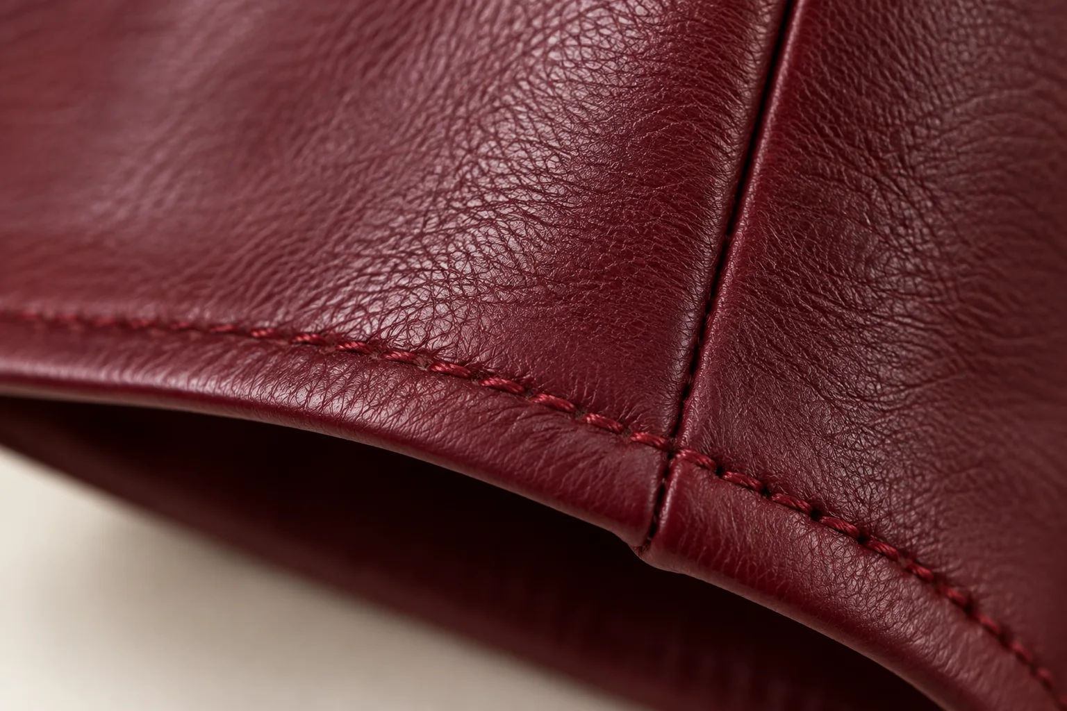 Burgundy long-cuff glove: extreme macro along its straight hem where tonal red topstitching follows thin turned leather edge and meets side seam.