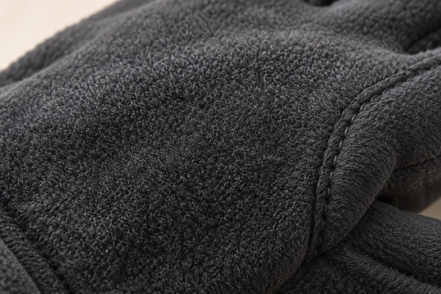 Charcoal fleece glove: macro of hand panel with soft short brushed pile, a small curved tonal finger-side seam at edge.